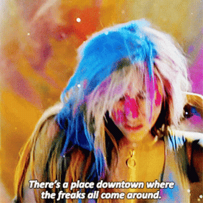 A Woman With Blue Hair And Pink Paint On Her Face Says " There 'S A Place Downtown Where The Freaks All Come Around " GIF
