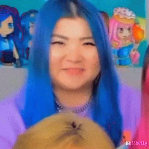 A Woman With Blue Hair Is Holding A Stuffed Animal In Front Of A Group Of Dolls . GIF
