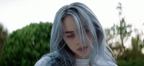 A Woman With Blue Hair Is Standing In Front Of A Bush With Her Eyes Closed And Her Hair Blowing In The Wind . GIF
