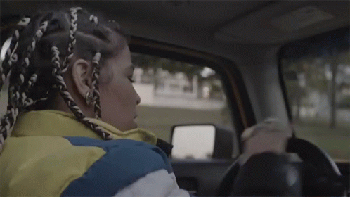 A Woman With Braids Is Driving A Car While Wearing A Yellow Jacket . GIF