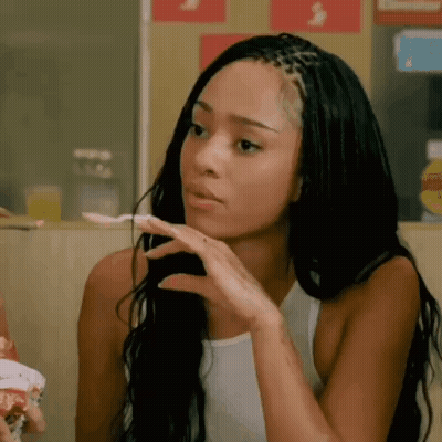 A Woman With Braids Is Sitting At A Table With A Spoon In Her Hand And Making A Funny Face . GIF