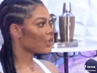 A Woman With Braids Is Sitting In Front Of A Table With Bottles Of Makeup On It . GIF