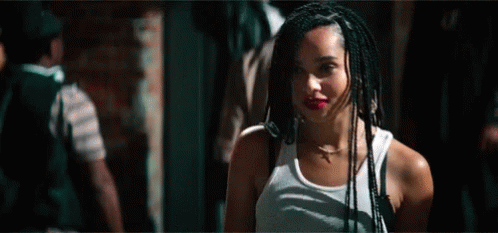A Woman With Braids Is Wearing A White Tank Top And Standing In A Dark Room . GIF