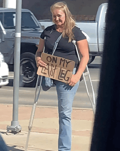 A Woman With Crutches Holding A Sign That Says On My Left Leg GIF