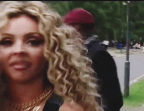 A Woman With Curly Blonde Hair Is Standing Next To A Man On A Sidewalk . GIF