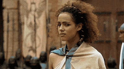 A Woman With Curly Hair And A Scarf Around Her Neck Is Standing In Front Of A Building . GIF
