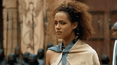 A Woman With Curly Hair And A Scarf Around Her Neck Is Standing In Front Of A Door . GIF