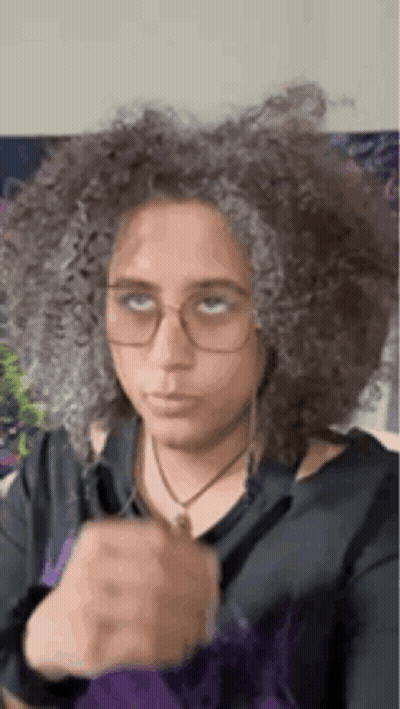 A Woman With Curly Hair And Glasses Is Wearing A Black Shirt And A Purple Jacket . GIF
