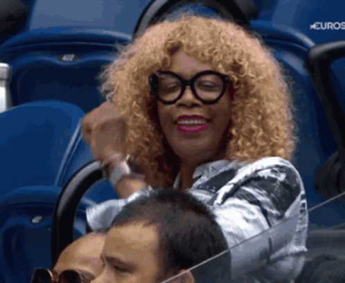 A Woman With Curly Hair And Glasses Sits In A Stadium With A Sign That Says Euros GIF