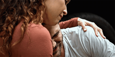 A Woman With Curly Hair Is Hugging A Man With His Eyes Closed GIF