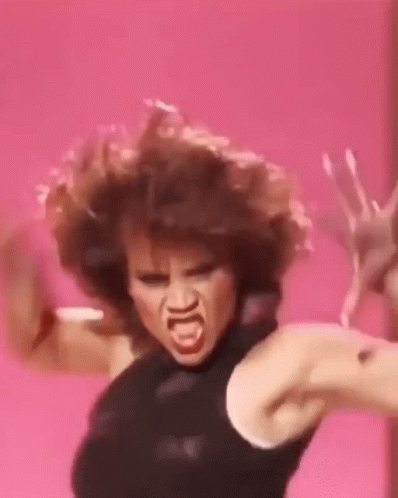 A Woman With Curly Hair Is Making A Funny Face While Dancing Against A Pink Background . GIF