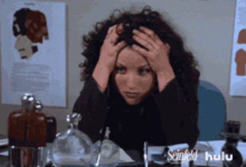 A Woman With Curly Hair Is Sitting At A Desk With Her Hands On Her Head And A Hulu Logo In The Corner GIF