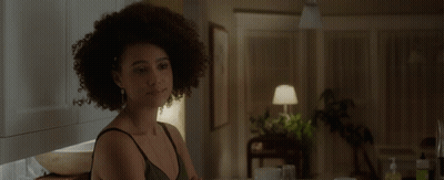 A Woman With Curly Hair Is Sitting At A Table In A Dark Room GIF