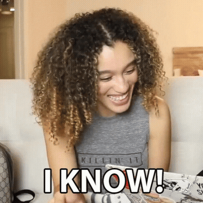 A Woman With Curly Hair Is Sitting On A Couch And Smiling While Holding A Magazine And Says I Know GIF