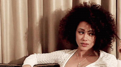 A Woman With Curly Hair Is Sitting On A Couch In Front Of A Curtain GIF