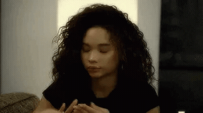 A Woman With Curly Hair Is Sitting On A Couch With Her Eyes Closed And Her Hands On Her Face . GIF