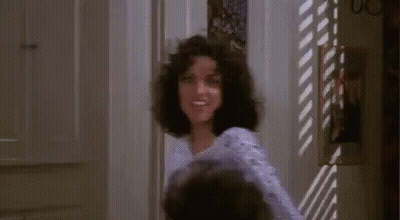 A Woman With Curly Hair Is Standing In A Doorway In A Room . GIF