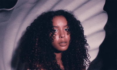 A Woman With Curly Hair Is Standing In Front Of A White Background GIF