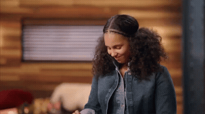 A Woman With Curly Hair Is Standing In Front Of A Wooden Wall In A Cabin And Smiling . GIF
