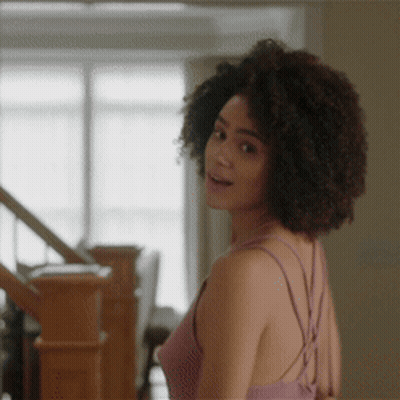 A Woman With Curly Hair Is Standing Next To A Staircase GIF