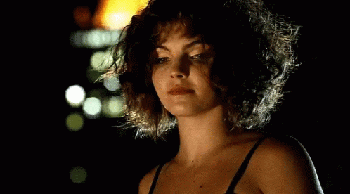A Woman With Curly Hair Is Wearing A Black Bra And Looking At The Camera In The Dark . GIF