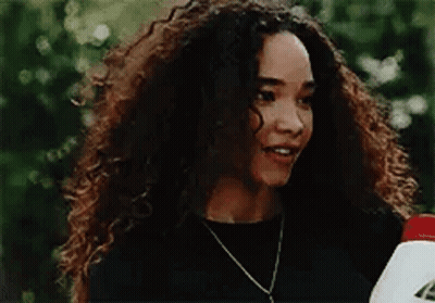 A Woman With Curly Hair Is Wearing A Black Shirt And A Necklace And Holding A Bottle . GIF