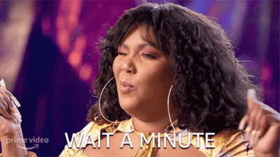 A Woman With Curly Hair Is Wearing A Gold Shirt And Earrings And Says Wait A Minute GIF