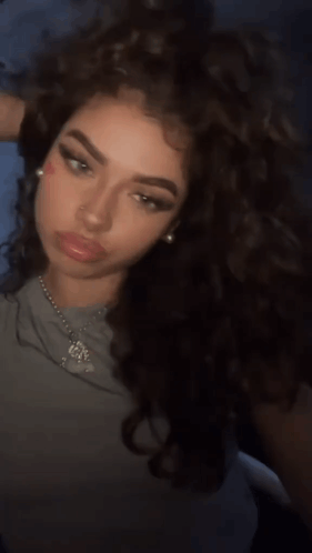 A Woman With Curly Hair Is Wearing A Necklace With A Pendant That Says ' Jesus ' On It GIF