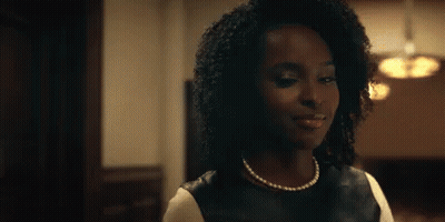 A Woman With Curly Hair Is Wearing A Pearl Necklace And A Black Leather Top . GIF