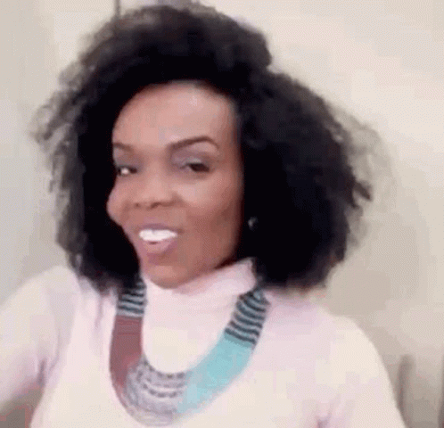 A Woman With Curly Hair Is Wearing A Pink Turtleneck And A Blue Necklace . GIF