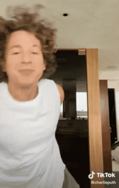 A Woman With Curly Hair Is Wearing A White Tank Top And Dancing In Front Of A Door . GIF