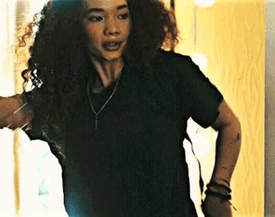 A Woman With Curly Hair Wearing A Black T-Shirt And A Necklace Is Standing In Front Of A Mirror . GIF