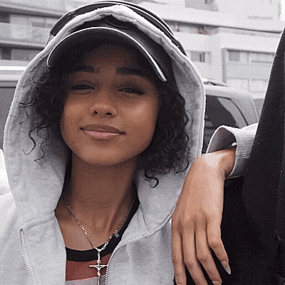 A Woman With Curly Hair Wearing A Hat And A Necklace With A Cross On It GIF