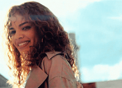 A Woman With Curly Hair Wearing A Trench Coat Is Smiling GIF