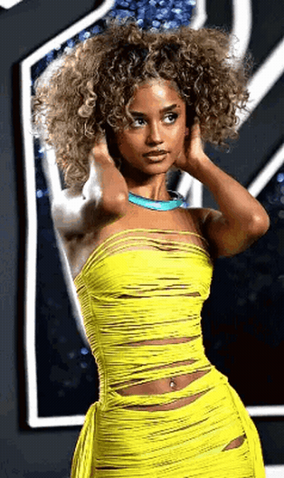 A Woman With Curly Hair Wearing A Yellow Dress And A Blue Necklace . GIF