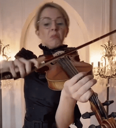 A Woman With Glasses Is Playing A Violin In Front Of A Chandelier GIF