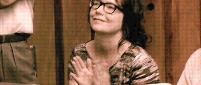 A Woman With Glasses Is Sitting At A Table With Her Hands Folded In Front Of Her Chest . GIF