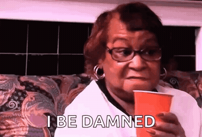 A Woman With Glasses Is Sitting On A Couch Holding A Red Cup And Saying `` I Be Damned '' . GIF