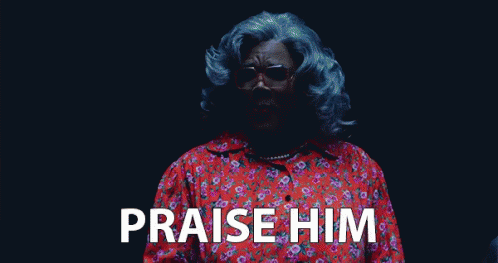 A Woman With Gray Hair And Glasses Is Standing In A Dark Room And Saying Praise Him . GIF
