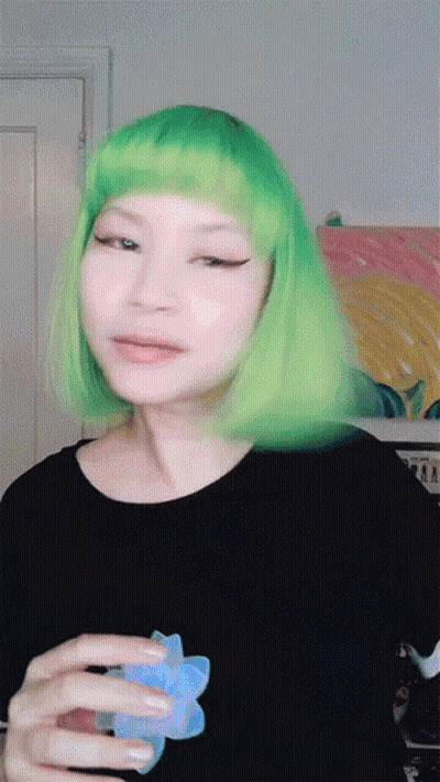 A Woman With Green Hair Is Holding A Blue Flower In Her Hand GIF