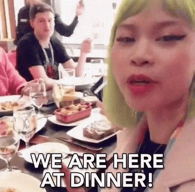 A Woman With Green Hair Is Sitting At A Table With Plates Of Food And Glasses Of Wine And Says We Are Here At Dinner . GIF
