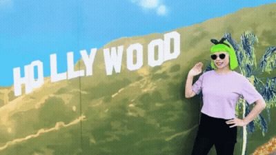 A Woman With Green Hair Is Standing In Front Of A Sign That Says Hollywood GIF