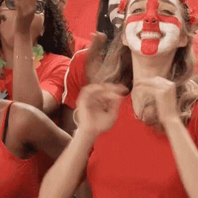 A Woman With Her Face Painted Red And White Is Sitting In A Stadium With Other People . GIF