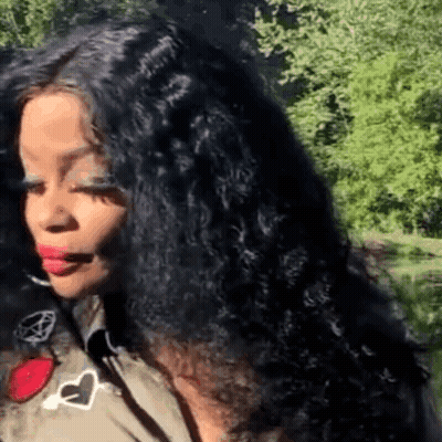 A Woman With Long Black Curly Hair Is Standing In Front Of Trees . GIF
