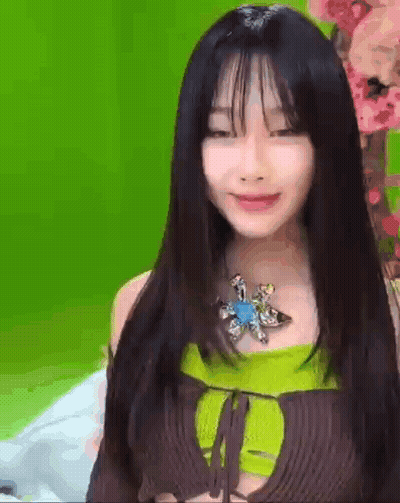 A Woman With Long Black Hair And Bangs Is Wearing A Green Top And A Brown Top . GIF