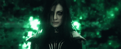 A Woman With Long Black Hair And Green Eyes Is Standing In Front Of A Forest . GIF