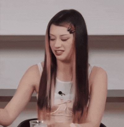 A Woman With Long Black Hair And Pink Streaks Is Sitting At A Table With A Glass Of Water . GIF