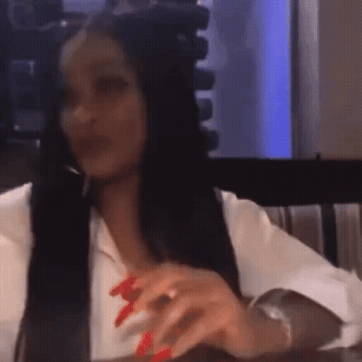 A Woman With Long Black Hair And Red Nails Is Sitting At A Table With Her Hands On The Table . GIF