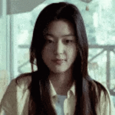 A Woman With Long Black Hair Is Sitting In Front Of A Window And Making A Funny Face . GIF