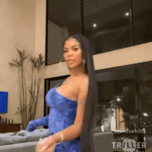 A Woman With Long Black Hair Is Standing In A Living Room Wearing A Blue Dress And Gloves . GIF